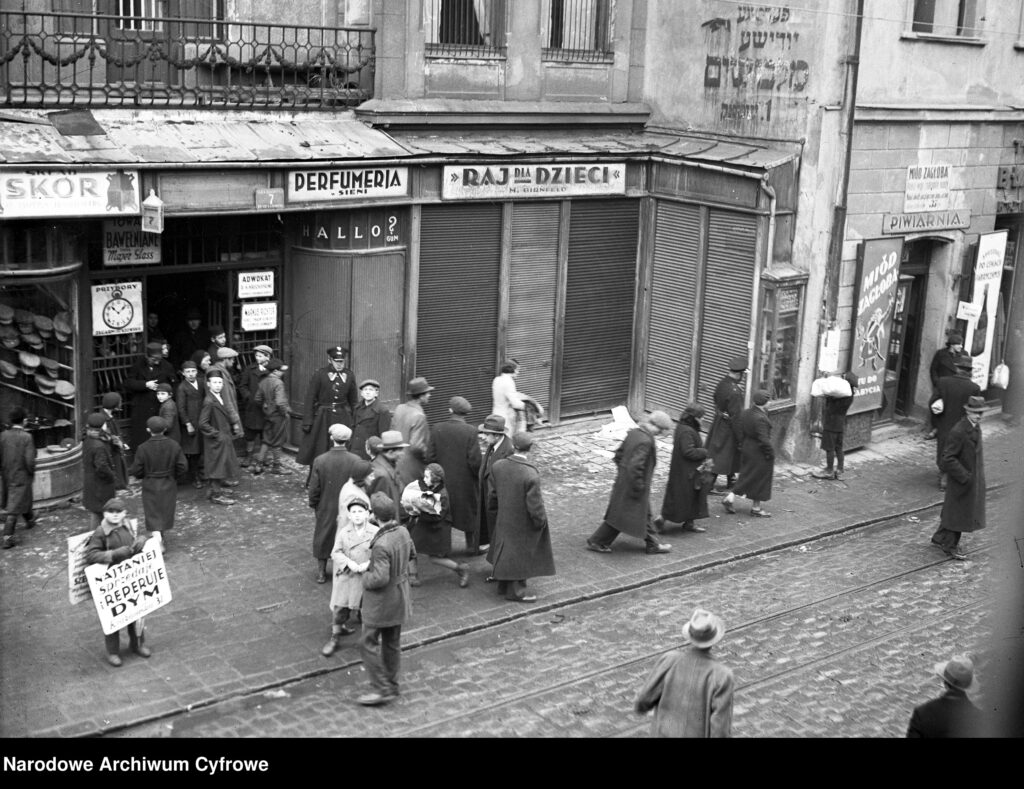 Pre-war Kraków street with historic tenement houses at numbers 5 and 7, showing shop signs in Polish and Jewish names, pedestrians, and a policeman.
